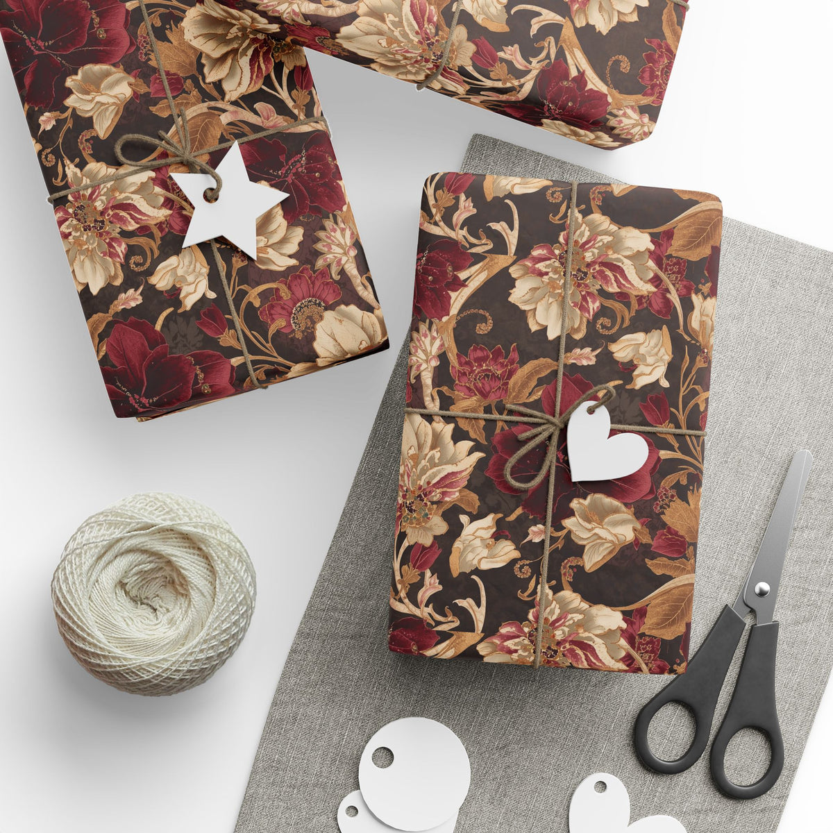Three gift boxes wrapped in a vintage floral patterned paper, tied with twine and decorated with white paper tags, arranged on a gray fabric surface with a ball of twine, scissors, and blank tags.