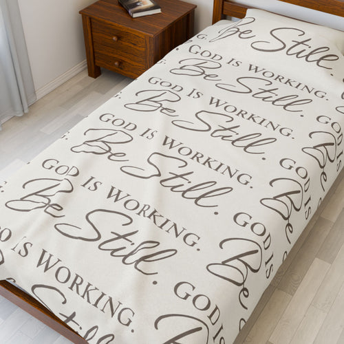 Blanket with 'Be Still' and 'God is Working' text on a bed in a bedroom.