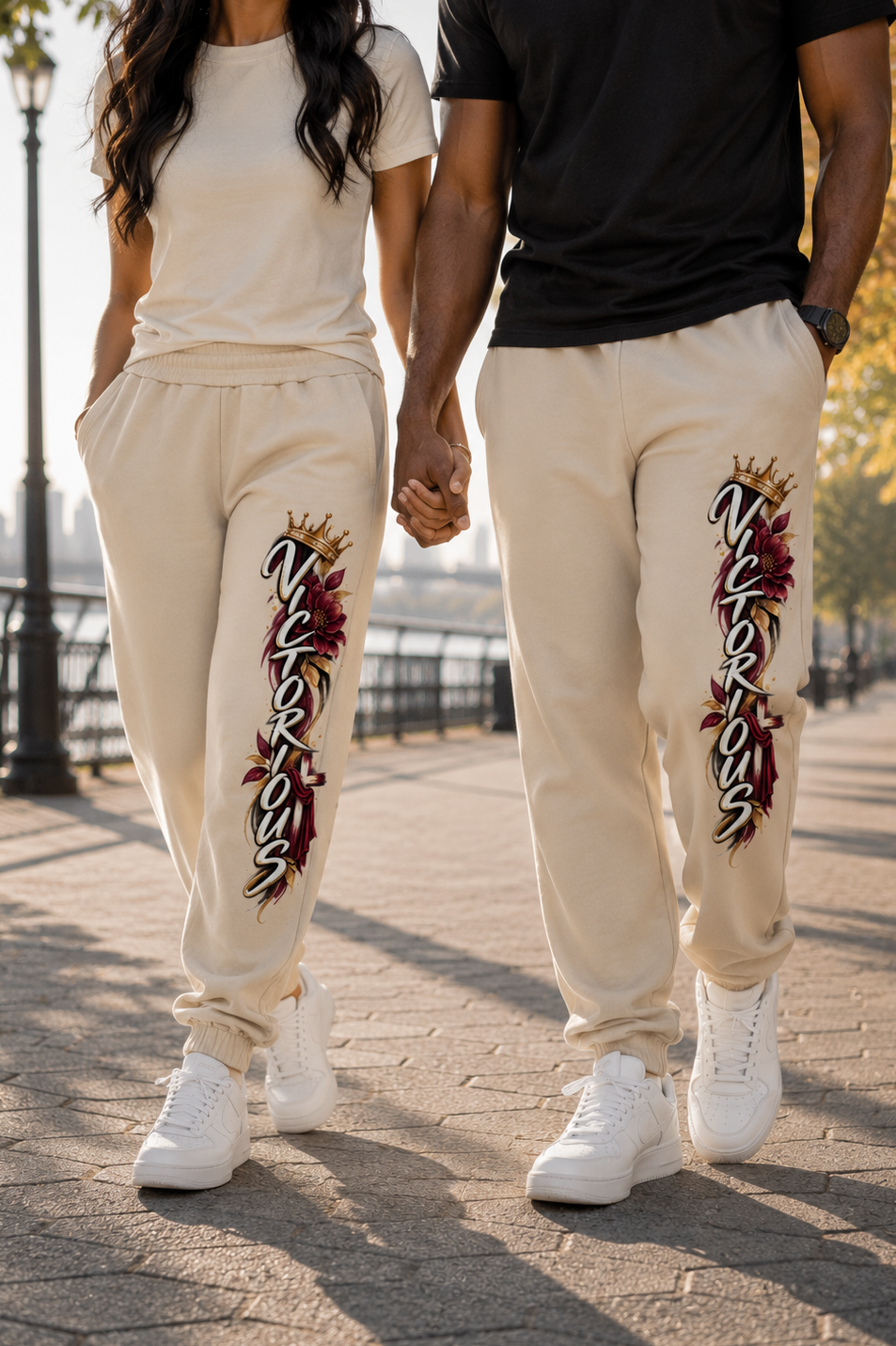 A couple walking hand-in-hand outdoors on a paved path near water. Both are wearing matching cream sweatpants with a large vertical graphic on the left leg featuring the word 'Nikki' in stylized lettering with red and gold details.