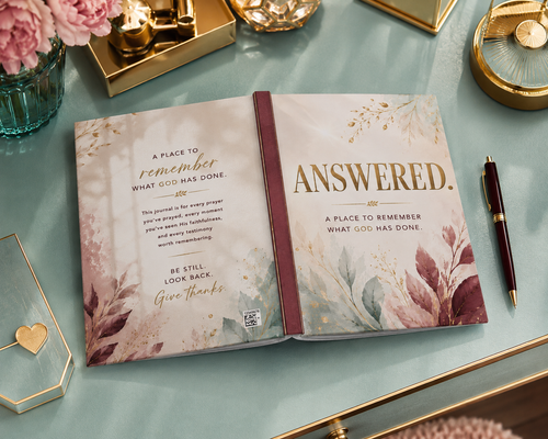 An open journal or prayer book with floral cover art and the title 'ANSWERED.' displayed on a light blue surface, surrounded by gold-toned decorative objects, a pen, and pink flowers.