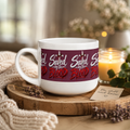 A white ceramic mug with a dark red band around the middle featuring the text 'Saved by the Blood' in white and red gothic-style lettering, styled on a wooden tray with a candle, knit blanket, and dried flowers.